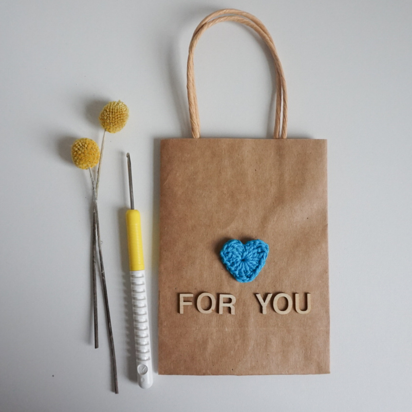 Tüte aus Kraftpapier mit einem gehäkelten Herz und dem Schriftzug "For You" aus Holzbuchstaben